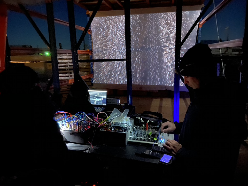 A person with a modular synthesizer setup performing in a dark space with a large visual projected behind them.
