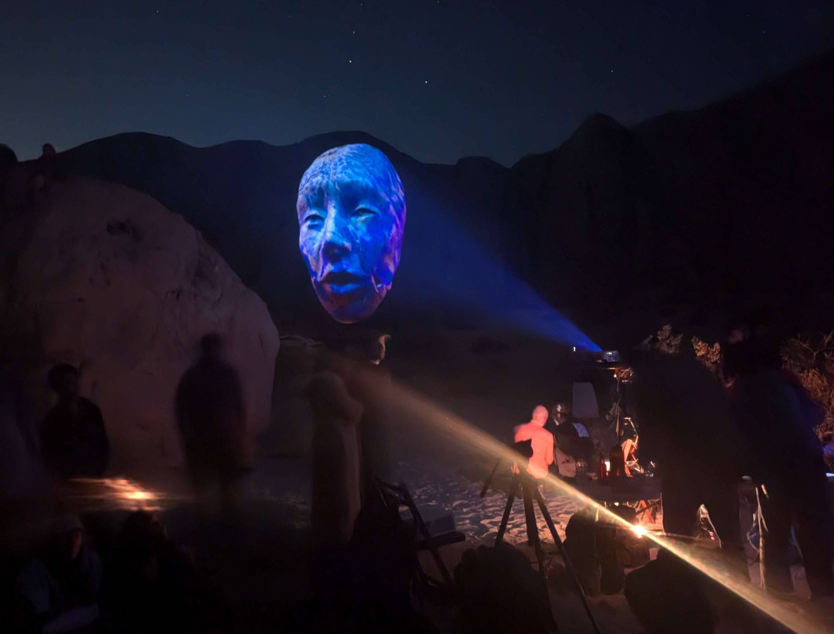 A large face projected onto a rock formation at night with an audience watching.
