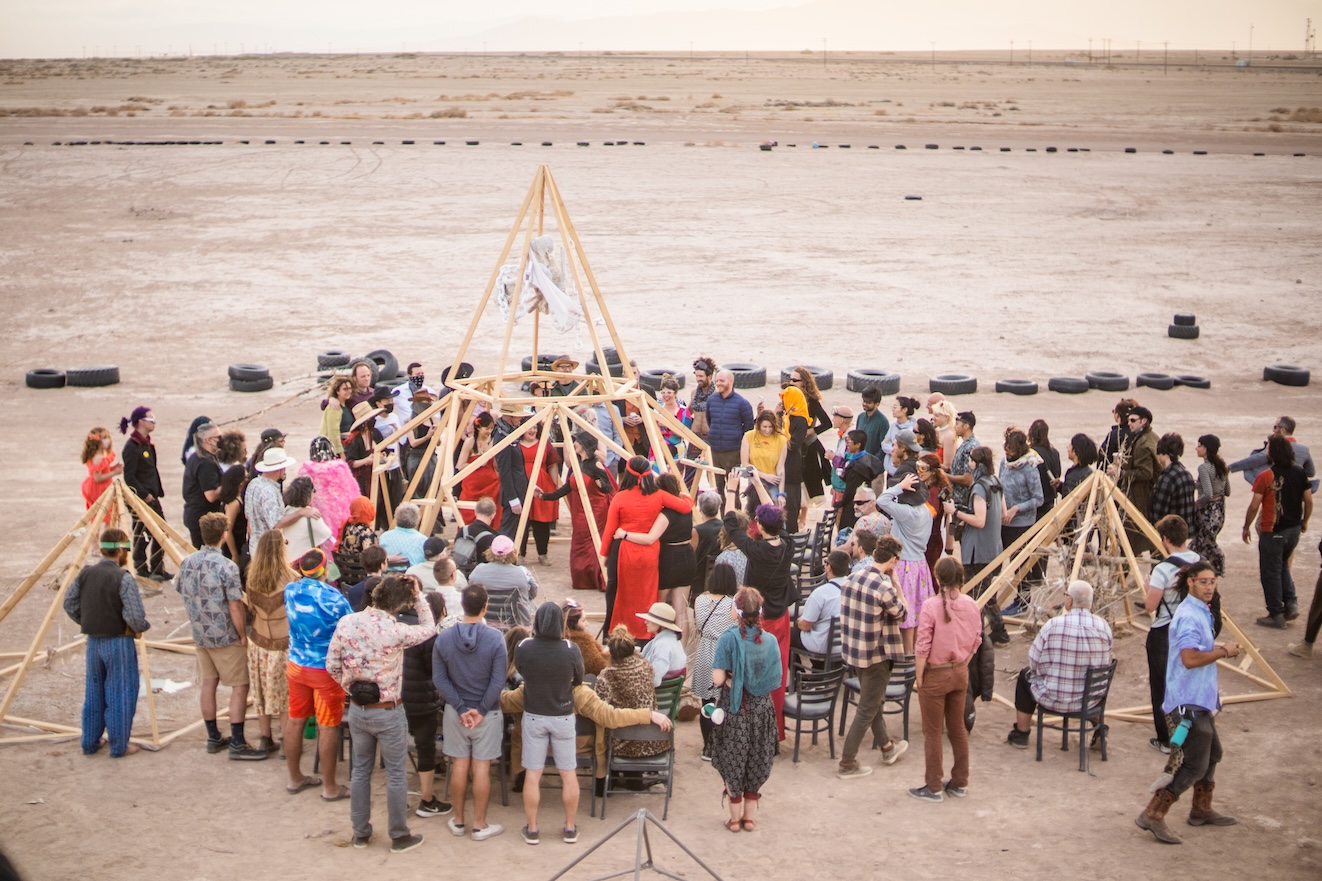 A large community gathering in a circle around wooden pyramid structures.